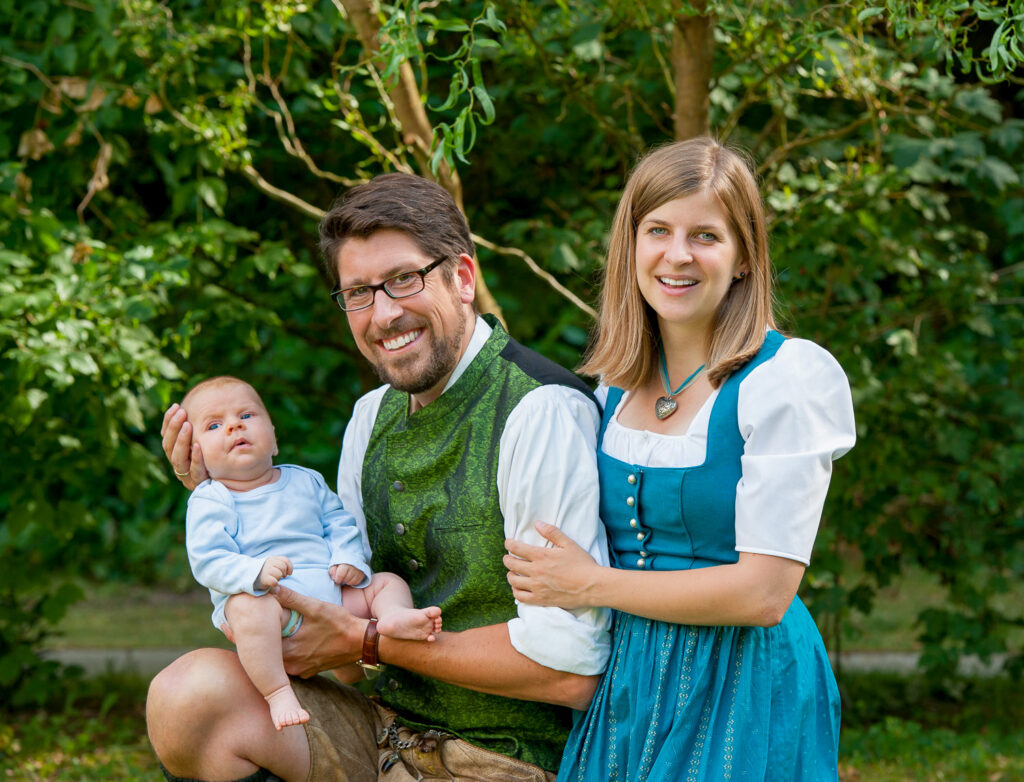 Hochzeitsfotograf Erlangen-Eine junge Familie ist in Trachten gekleidet in einem Park. Der Vater hält sein Baby behutsam mit einem glücklichen Lachen auf dem Arm-Coverbild Preise