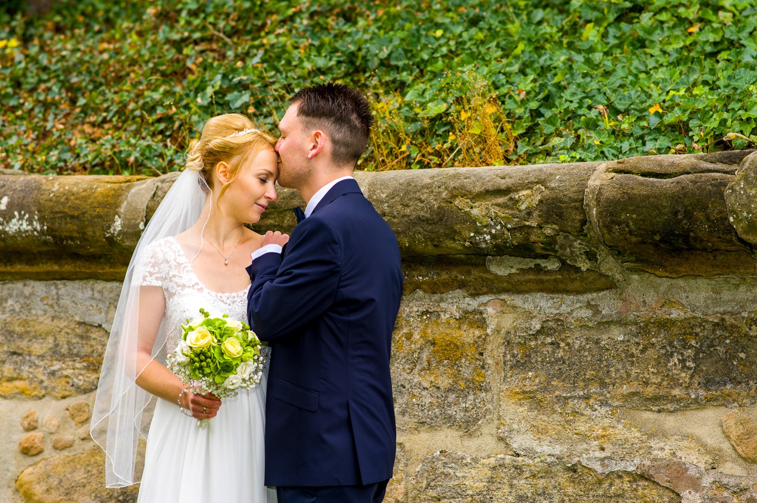 Hochzeitsfotograf Erlangen-Der Bräutigam küsst seine Braut liebevoll auf die Stirn. Im Hintergrund ist eine alte Steinmauer und etwas grün zu sehen